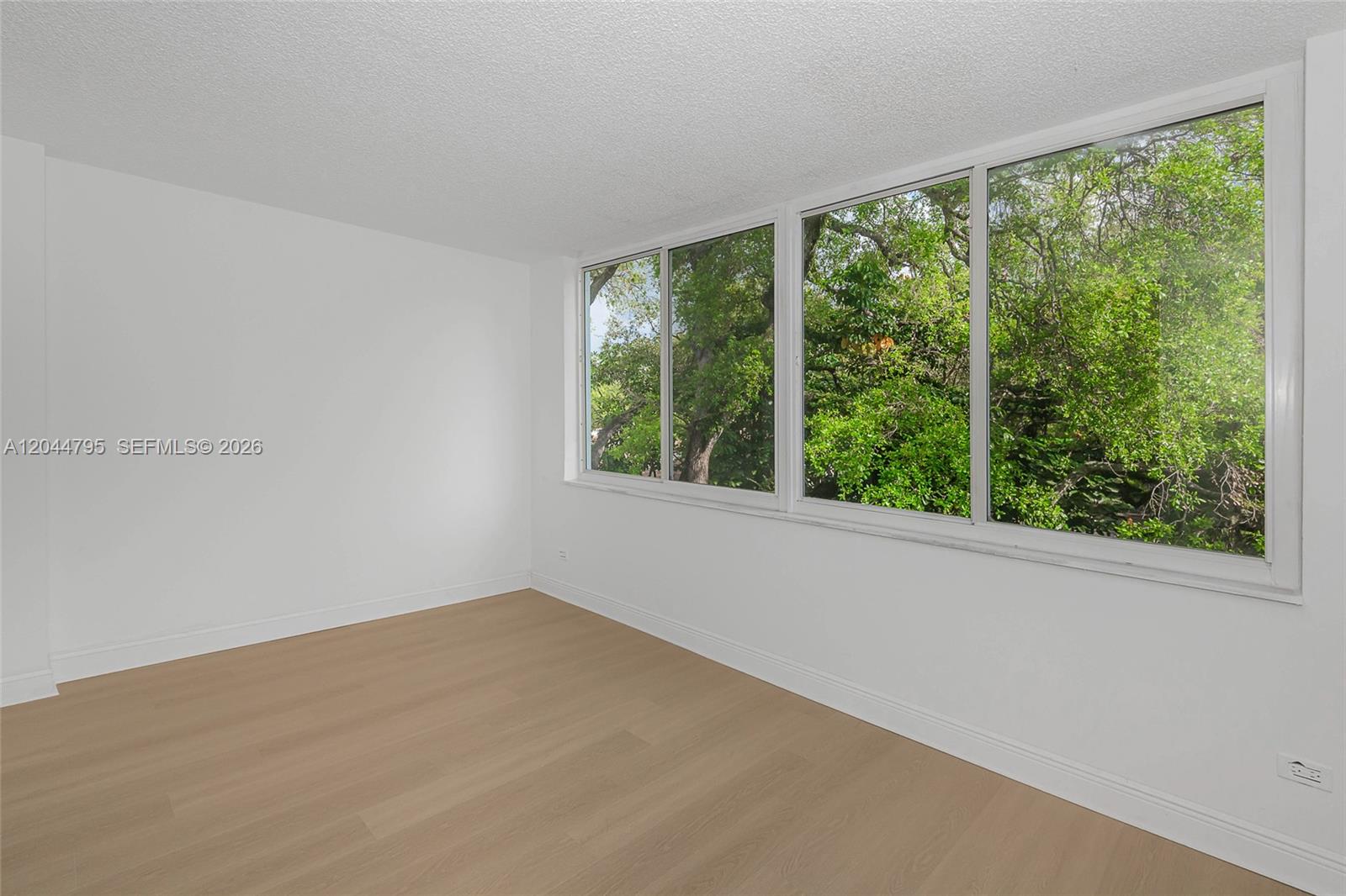 An empty room features light wood-look flooring, white walls, and a large window overlooking trees — 445 NE 195th St # 326, Miami FL 33179, MIAMI, FL