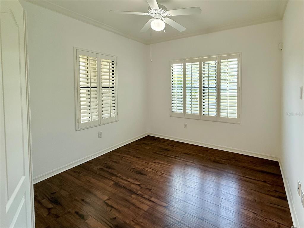An empty room features white walls, dark wood-look flooring, a ceiling fan, and plantation shutters — 10735 TROPHY DRIVE, ENGLEWOOD, FL
