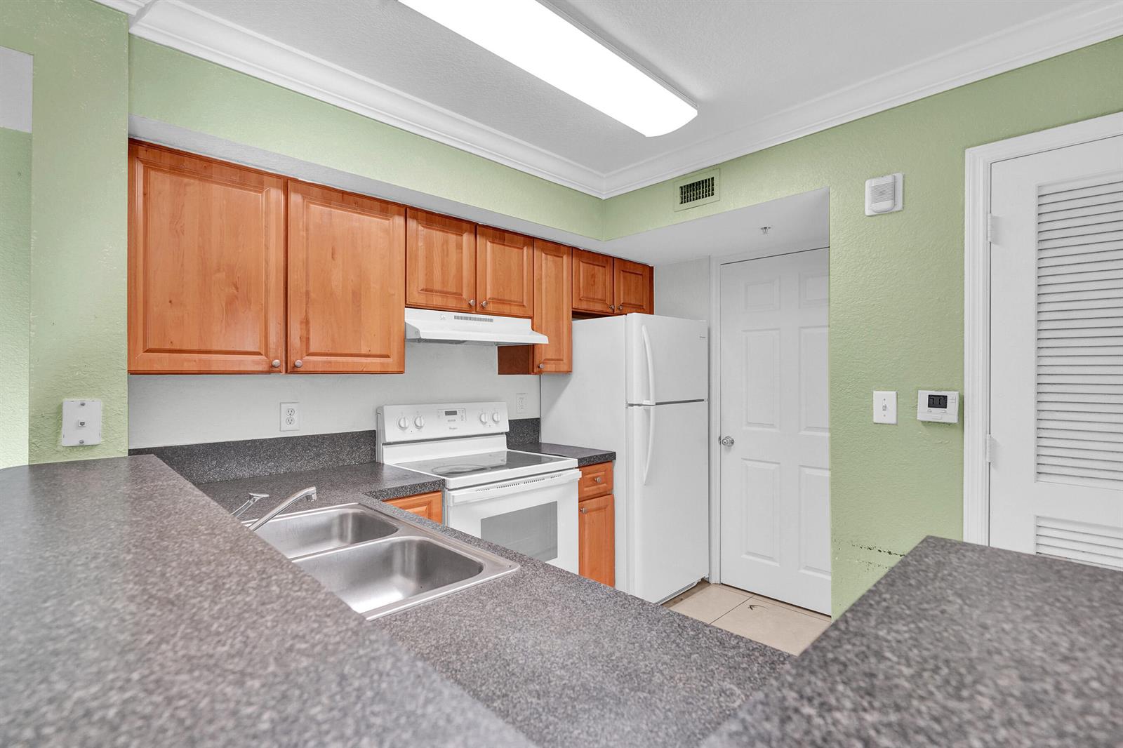 A kitchen features light green walls, wood cabinets, dark countertops, and white appliances — 15075 Michelangelo Boulevard # 103, Delray Beach FL 33446, DELRAY BEACH, FL