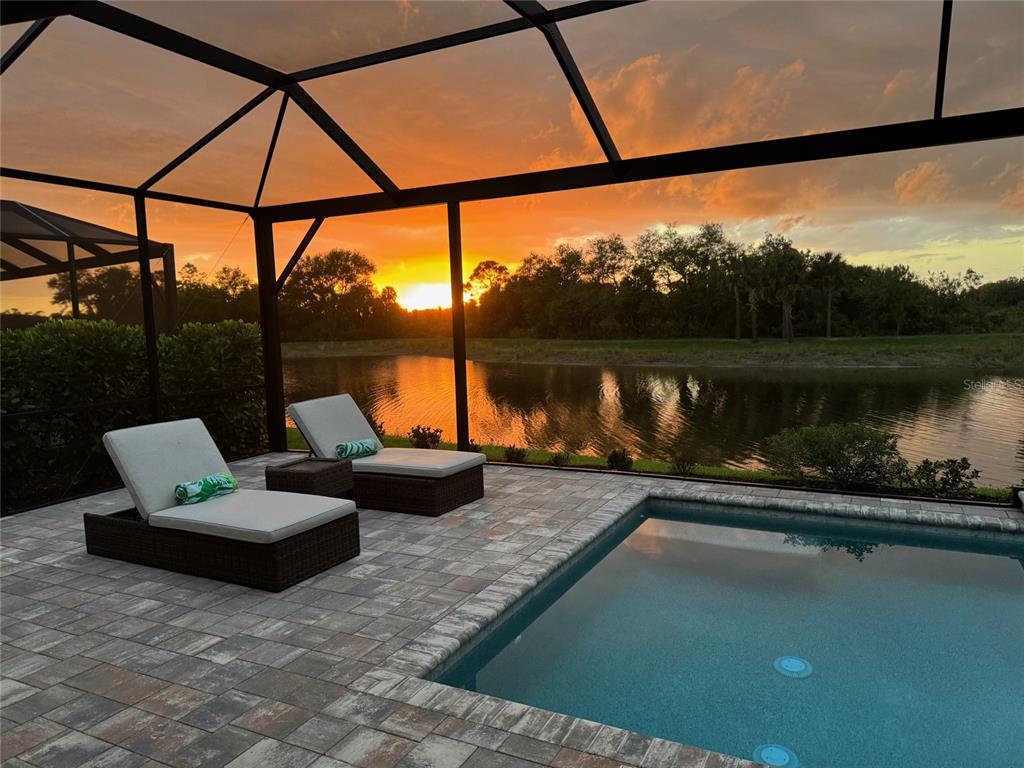 A screened-in patio features a swimming pool and lounge chairs overlooking a sunset lake view — 18035 HOME RUN DRIVE, VENICE, FL
