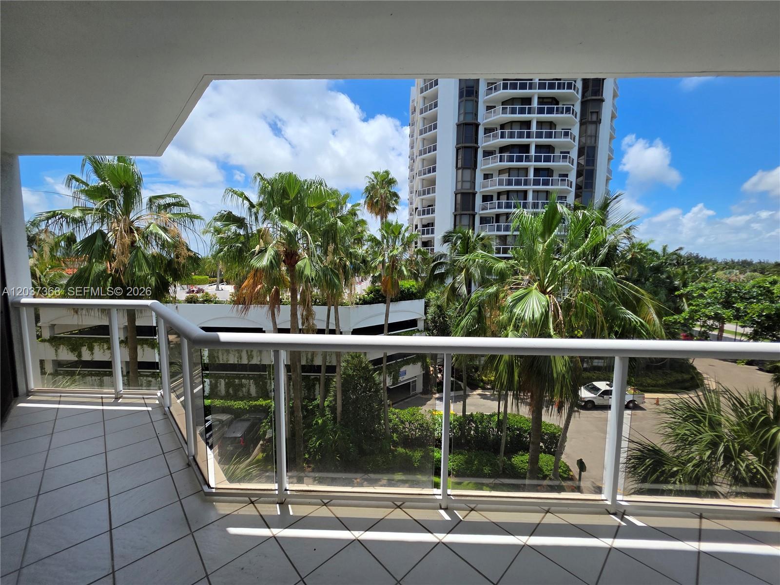 A balcony with white tile flooring overlooks palm trees, a parking structure, and a high-rise building — AVENTURA, FL