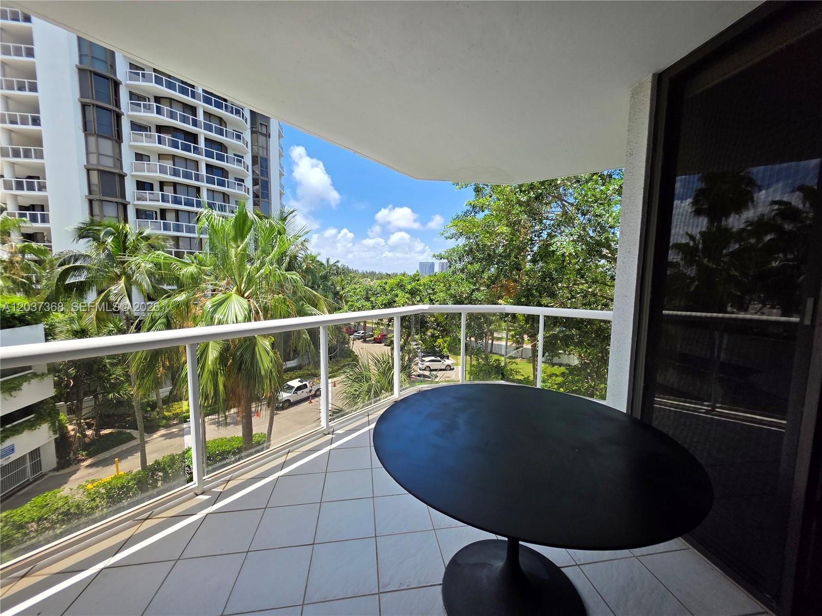 A balcony with a round black table overlooks palm trees and a neighboring apartment building — AVENTURA, FL