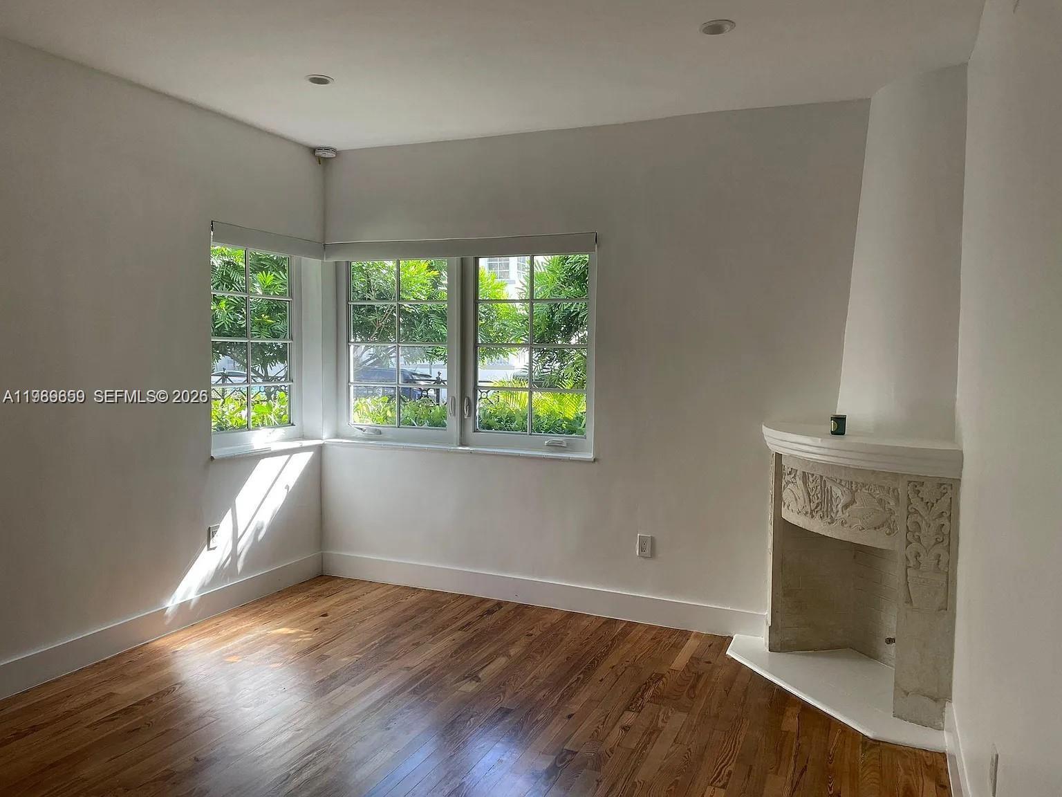 An empty room features hardwood floors, white walls, corner windows, and a decorative stone fireplace — 727 14th Pl # 1, Miami Beach FL 33139, MIAMI BEACH, FL