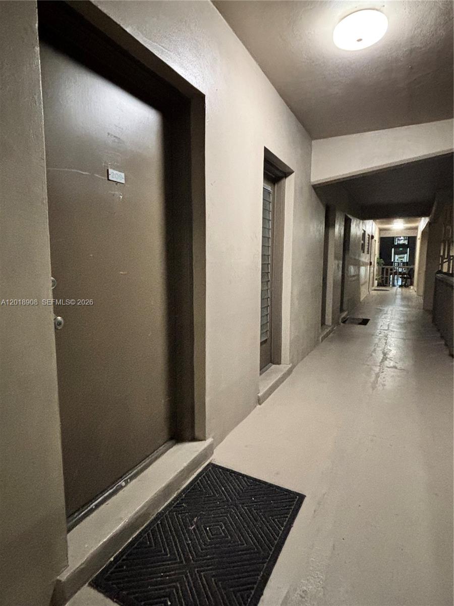 A brown apartment door with a black floor mat sits in a concrete hallway — 1480 E Sample Rd # 209, Pompano Beach FL 33064, POMPANO BEACH, FL