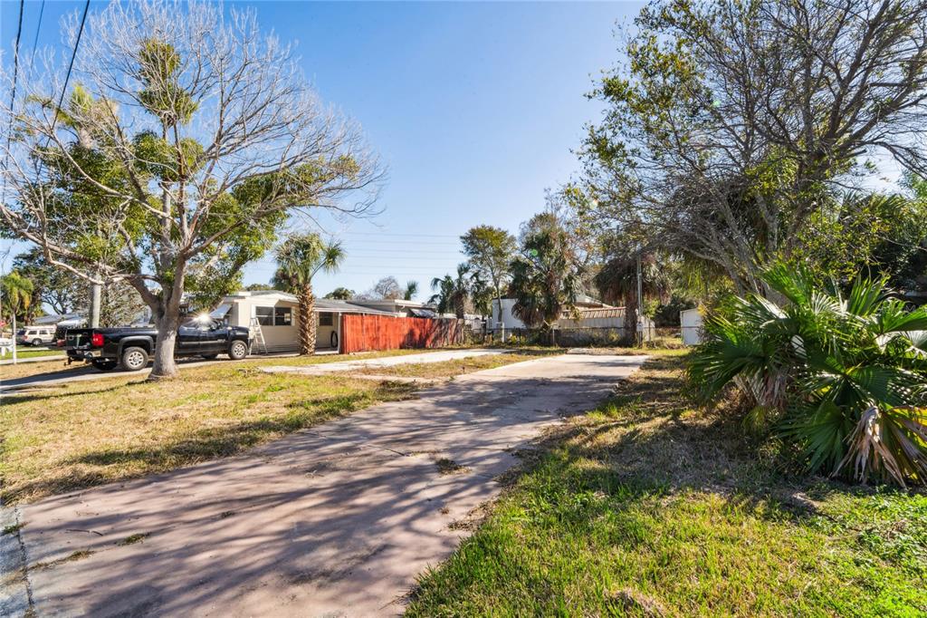 A concrete driveway leads toward a mobile home with a red fence and surrounding trees — 736 DRIFTWOOD LANE, TARPON SPRINGS, FL