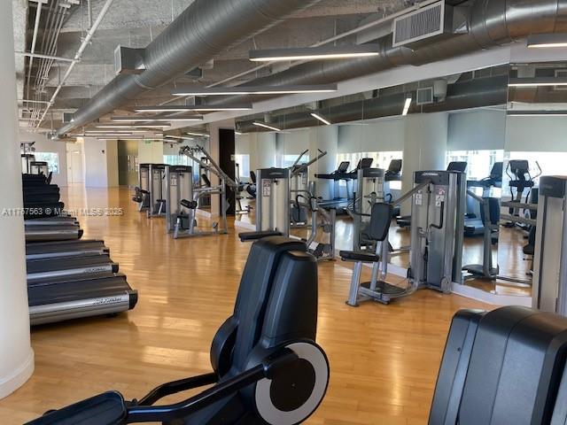 A gym features rows of exercise equipment, a light wood floor, and exposed industrial ceiling ducts — 100 SE 2 ST # 2000, Miami FL 33131, MIAMI, FL