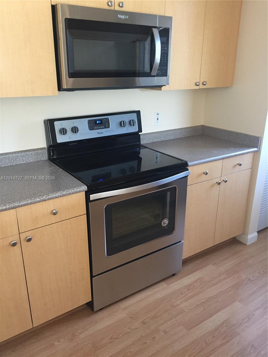A galley kitchen features light wood cabinets, gray countertops, stainless steel appliances, and wood flooring — 1900 Van Buren St # 214B, Hollywood FL 33020, HOLLYWOOD, FL