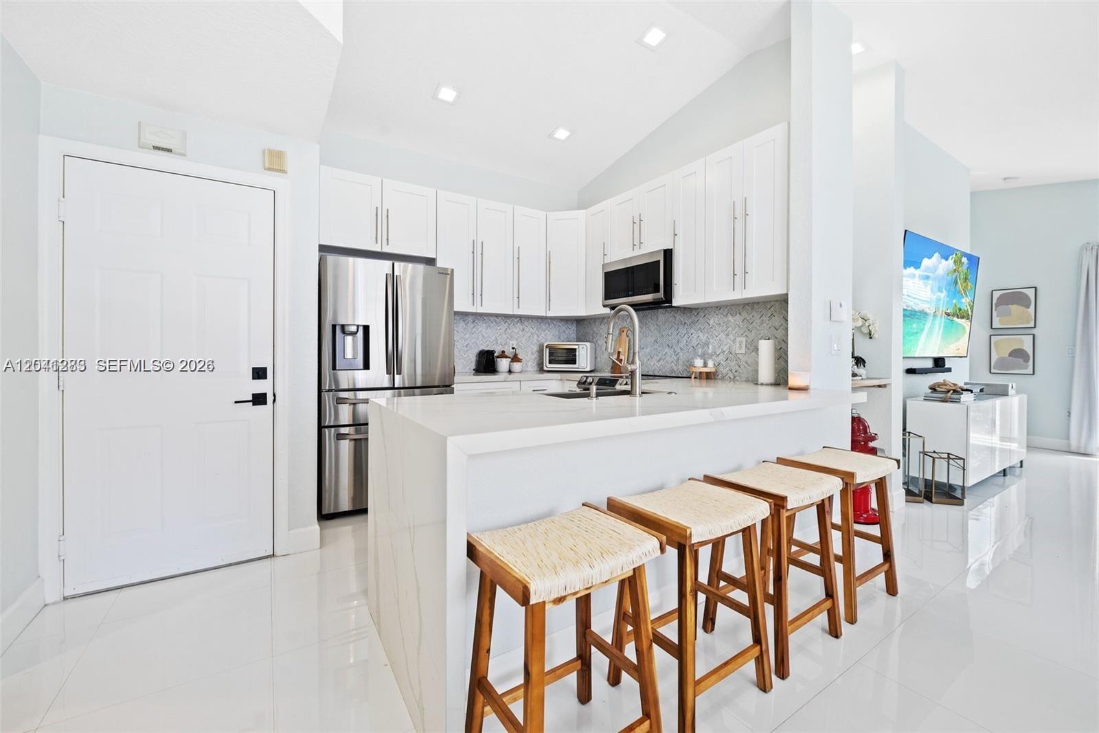 A modern kitchen features white cabinetry, stainless steel appliances, a waterfall island, and bar stools — 18755 SW 27th Ct, Miramar FL 33029, MIRAMAR, FL