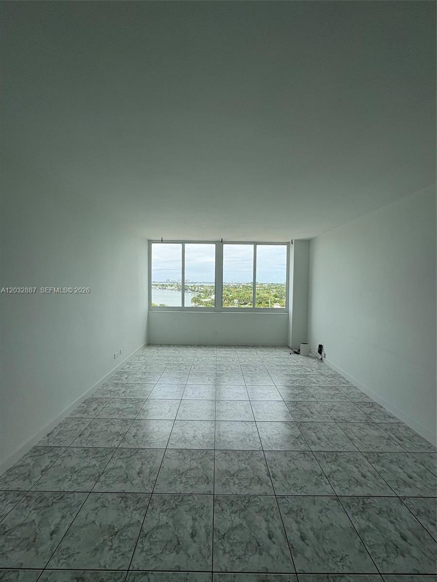 An empty room features white walls, light-colored marble-patterned tile flooring, and a large window — 7441 Wayne Ave # 8B, Miami Beach FL 33141, MIAMI BEACH, FL