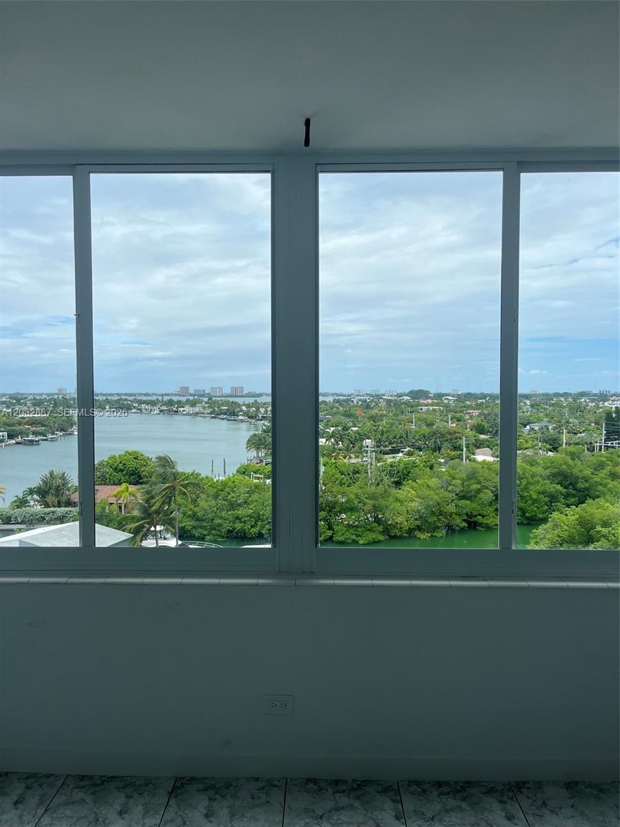 A room with large windows overlooking a bay, lush greenery, and distant buildings — 7441 Wayne Ave # 8B, Miami Beach FL 33141, MIAMI BEACH, FL