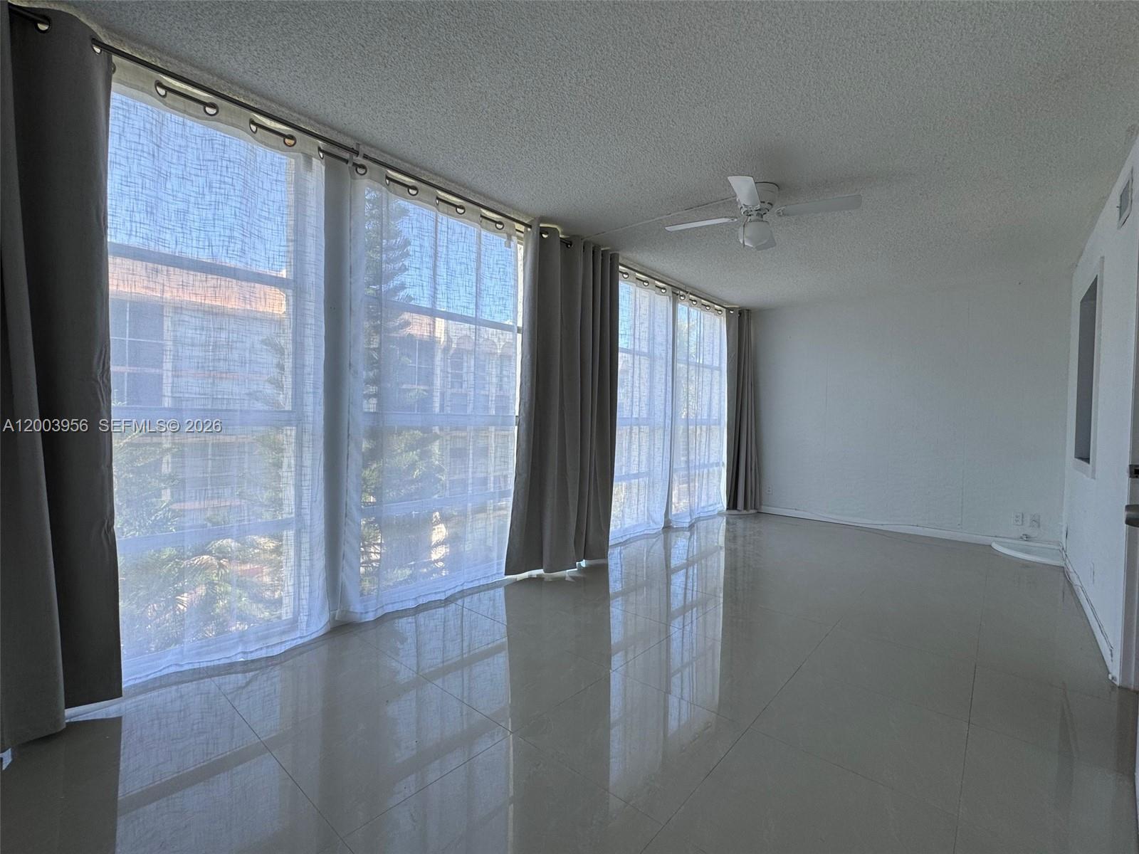 A bright, empty room features large windows with curtains, a ceiling fan, and tiled floors — 851 Three Islands Blvd # 411, Hallandale Beach FL 33009, HALLANDALE BEACH, FL