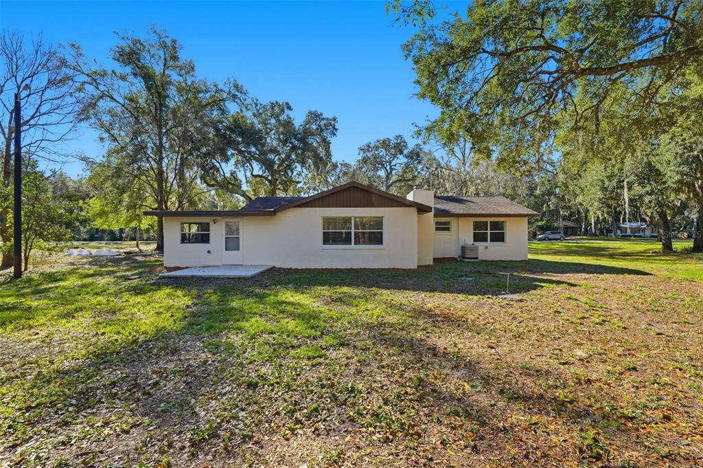 The back exterior of a single-story house features a concrete patio and large shade trees — 34071 MADISON AVENUE, DADE CITY, FL
