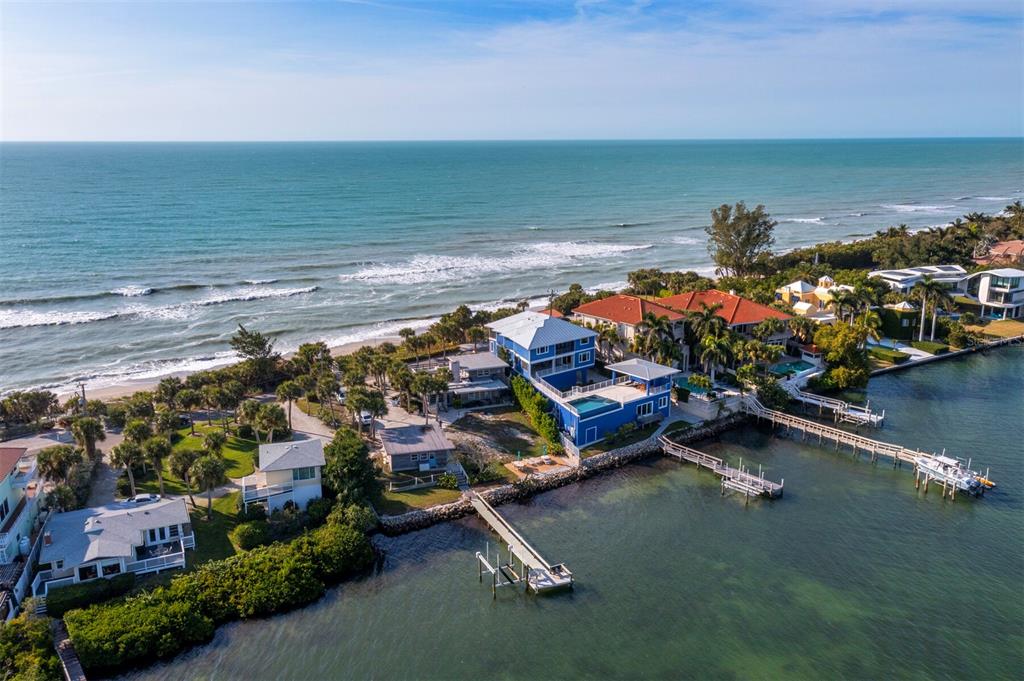An aerial view shows a blue multi-story house on a narrow strip of land between the ocean and a bay — 1620 CASEY KEY ROAD, NOKOMIS, FL