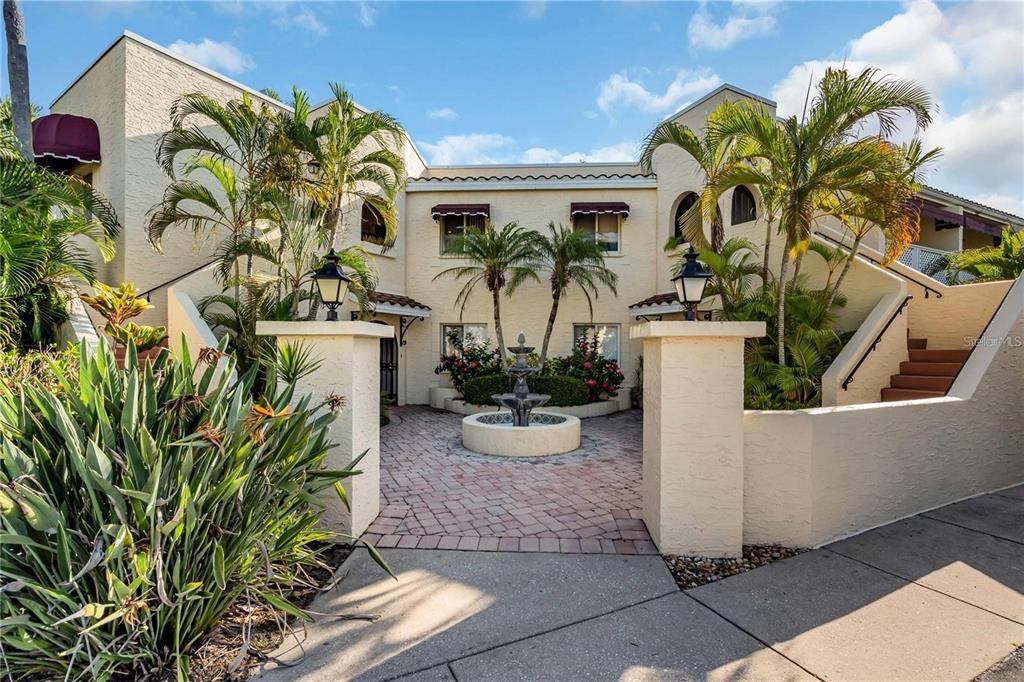 A courtyard with a fountain sits between two stucco buildings with palm trees and stairs — 6101 34TH ST W UNIT 27C, BRADENTON, FL