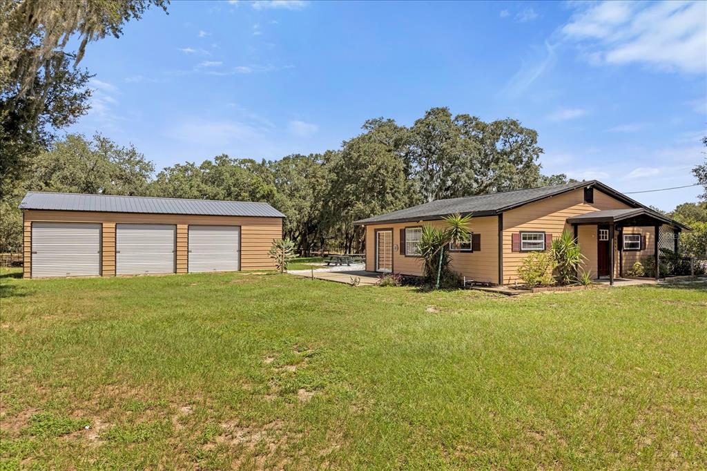 A tan house with a metal-roofed three-car garage sits on a grassy lot with trees — 14043 TENNESSEE AVENUE, ASTATULA, FL