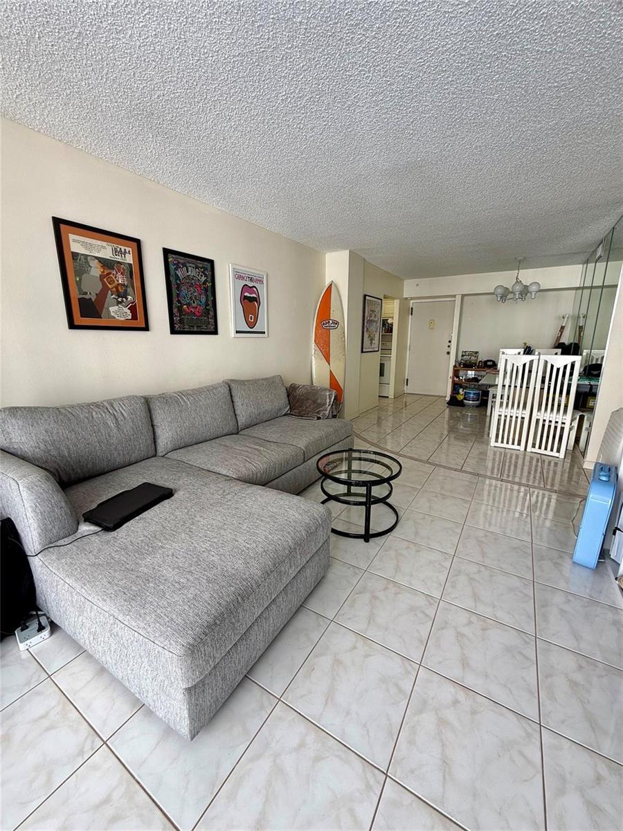 A living room with a grey sectional sofa, tiled floors, and a surfboard leaning against the wall — 111 Briny Avenue # 1405, Pompano Beach FL 33062, POMPANO BEACH, FL