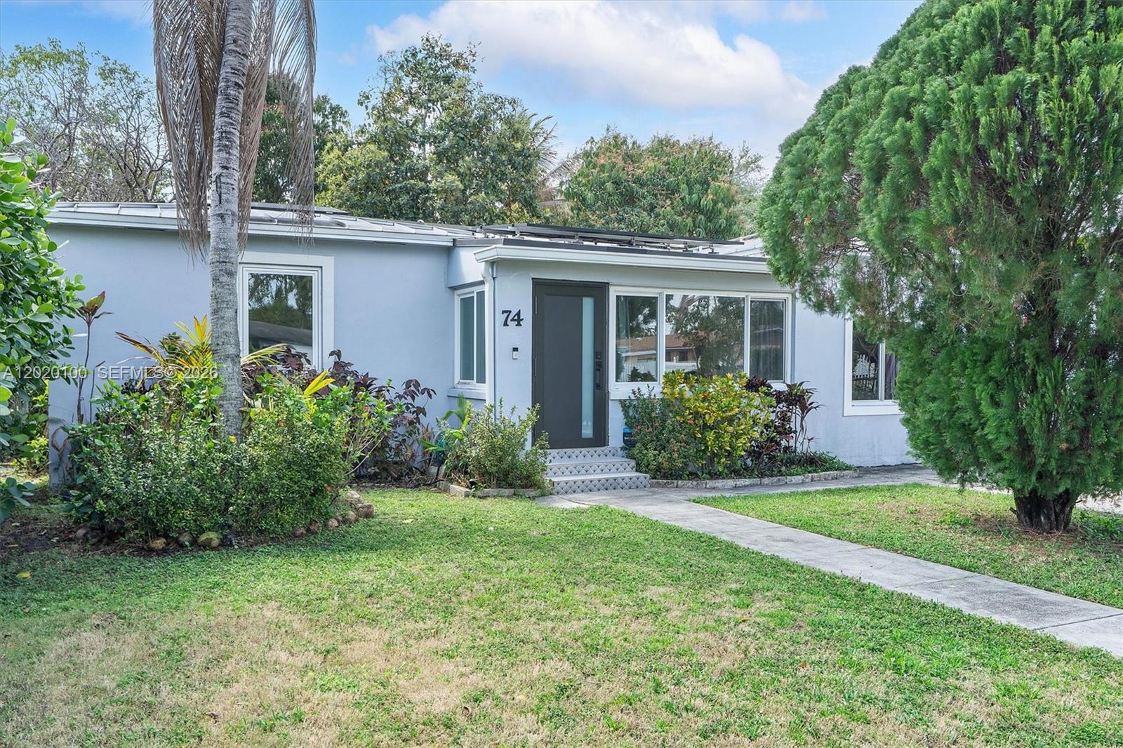 A light blue single-story house features a front door, large windows, and a landscaped yard — 74 NW 117th St, Miami FL 33168, MIAMI, FL
