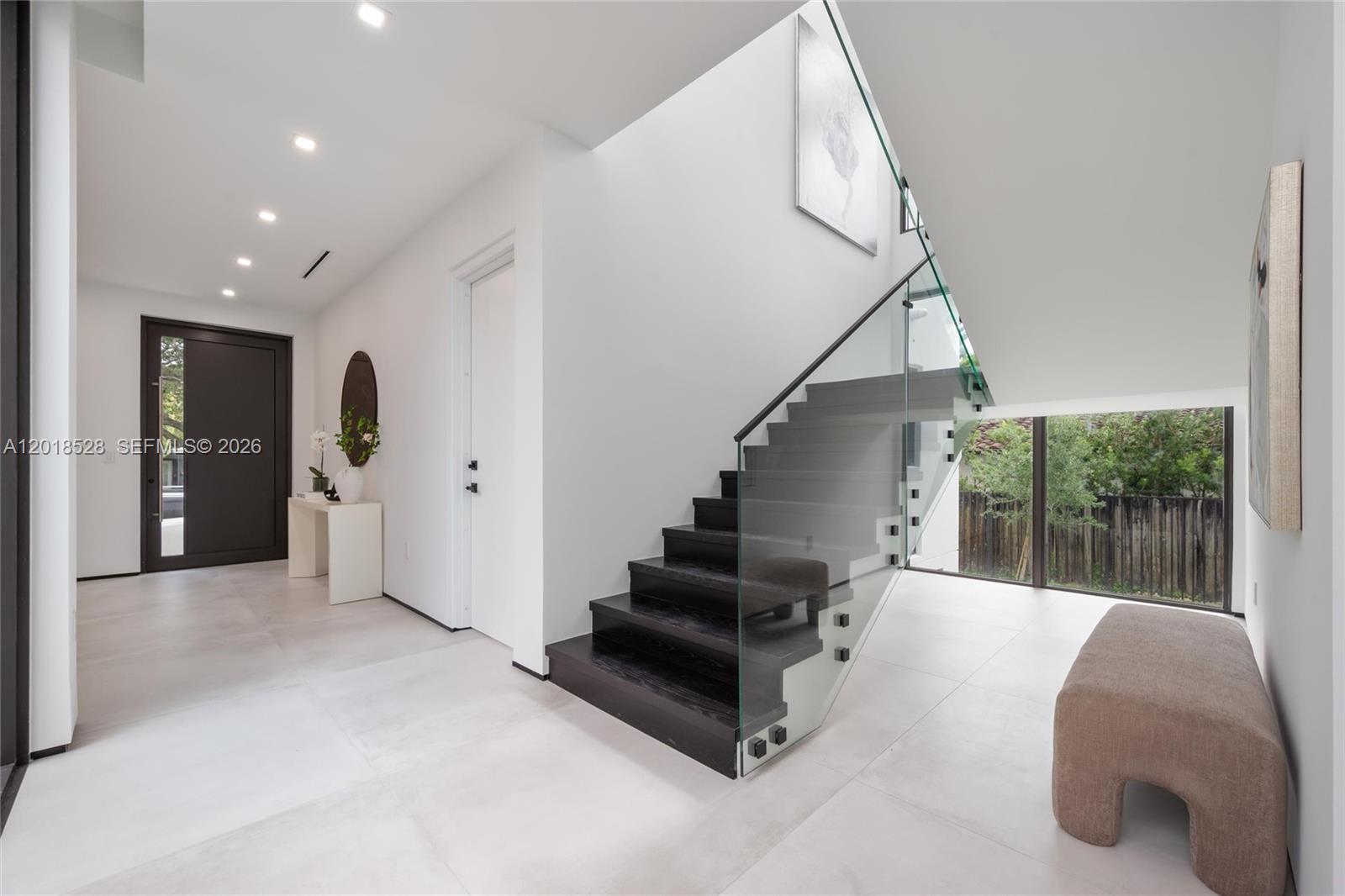 A modern entryway features a dark wood staircase with a glass railing and tiled floors — 2096 Alamanda Dr, North Miami FL 33181, NORTH MIAMI, FL