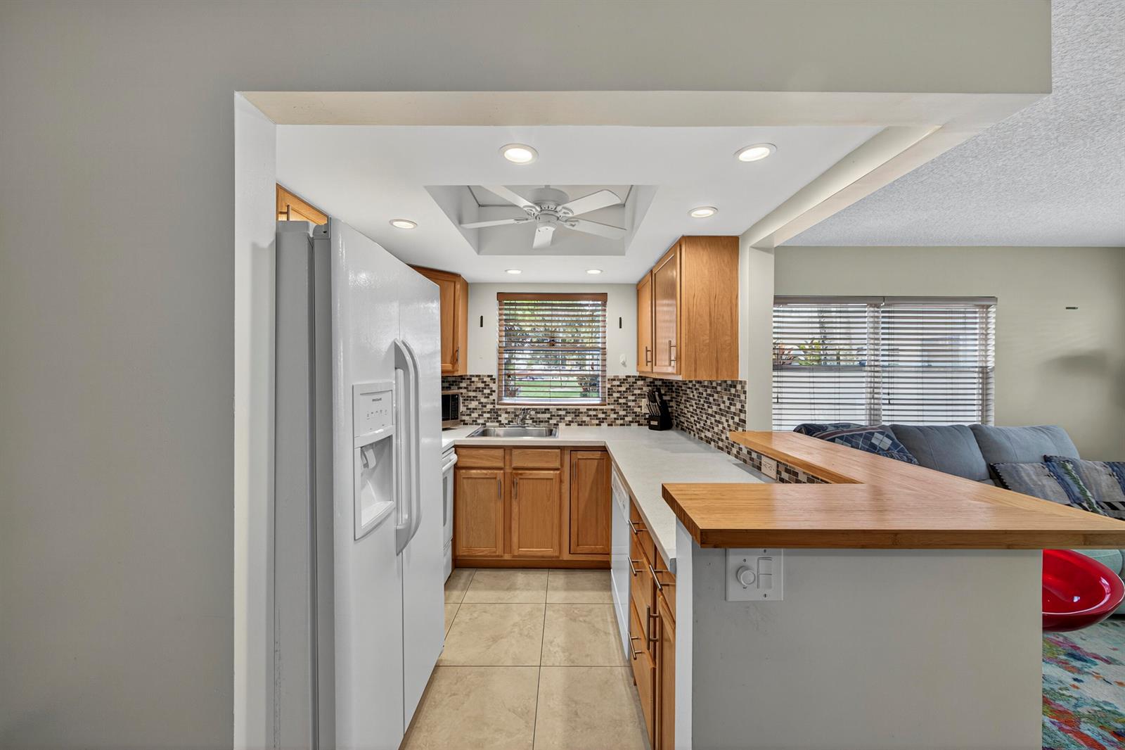 A kitchen features light wood cabinets, a tile backsplash, and a breakfast bar overlooking a living area — 125 Newport I, Deerfield Beach FL 33442, DEERFIELD BEACH, FL