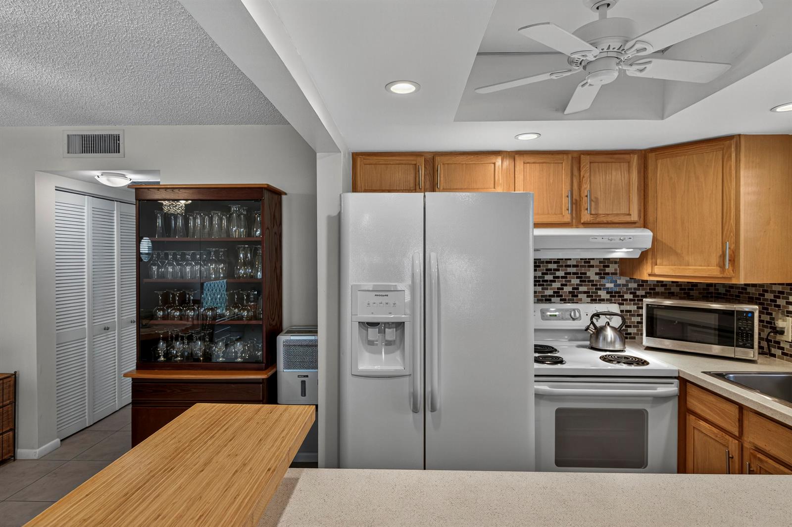 A kitchen features white appliances, wood cabinetry, a glass-front display cabinet, and a breakfast bar — 125 Newport I, Deerfield Beach FL 33442, DEERFIELD BEACH, FL