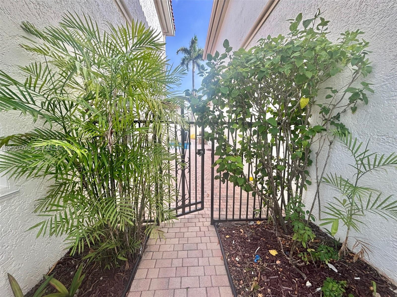 A paved walkway leads through a metal gate between two stucco walls with landscaping — 1141 Waterside Ln, Hollywood FL 33019, HOLLYWOOD, FL