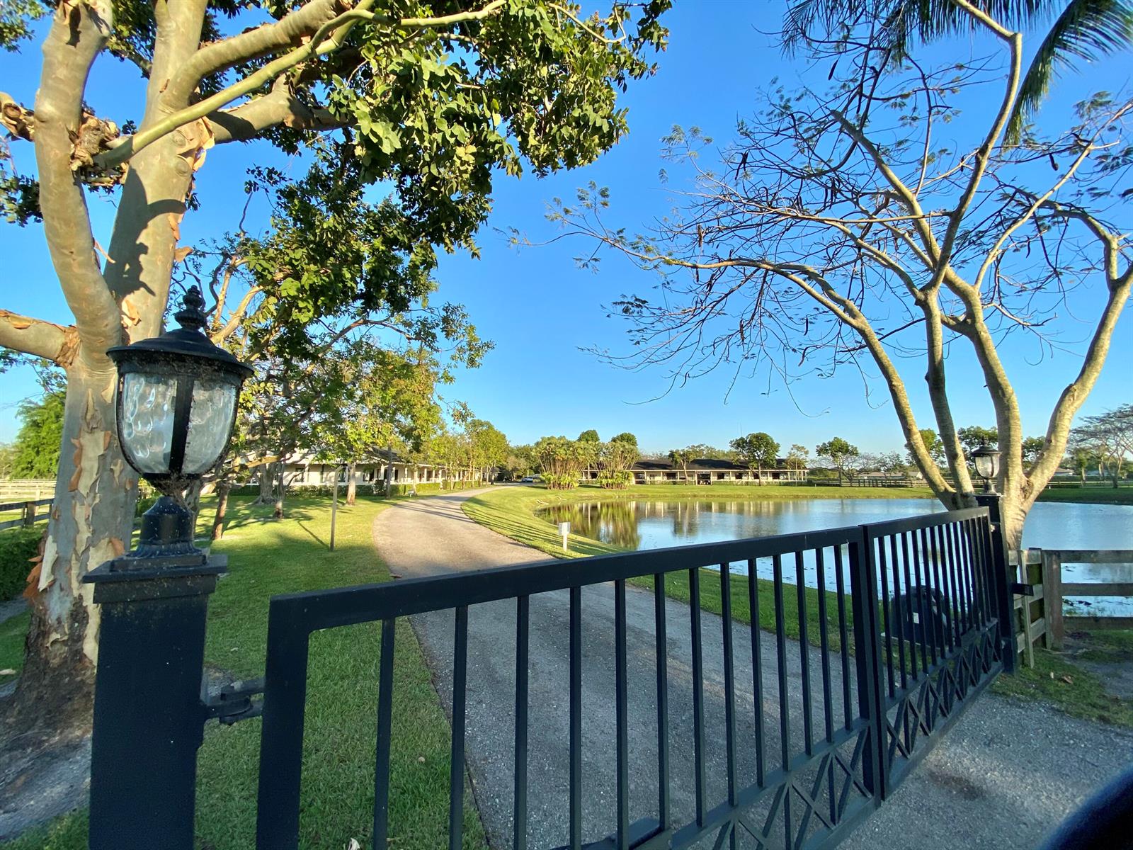 A black metal gate opens to a driveway leading toward a pond and distant buildings — 5110 S Shore Boulevard, Wellington FL 33449, WELLINGTON, FL
