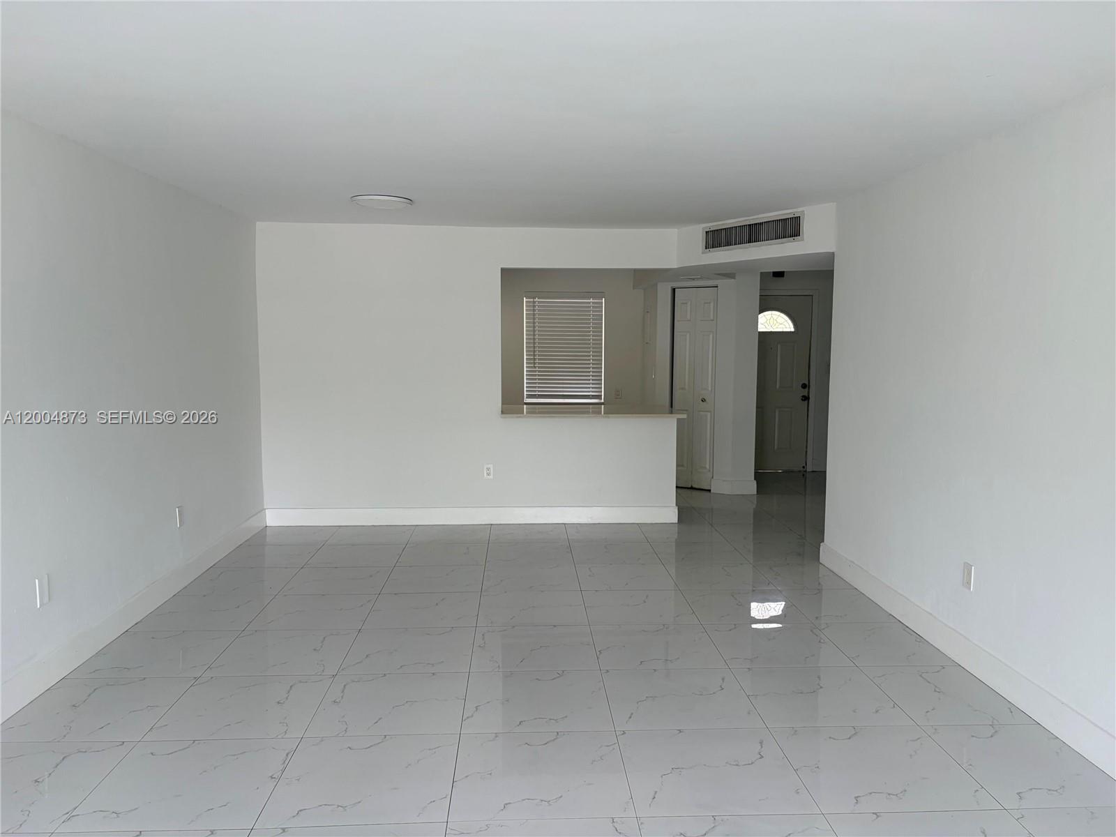 An empty room with white walls, large light-colored floor tiles, and a kitchen pass-through window — 1401 NE 191st St # D201, Miami FL 33179, MIAMI, FL