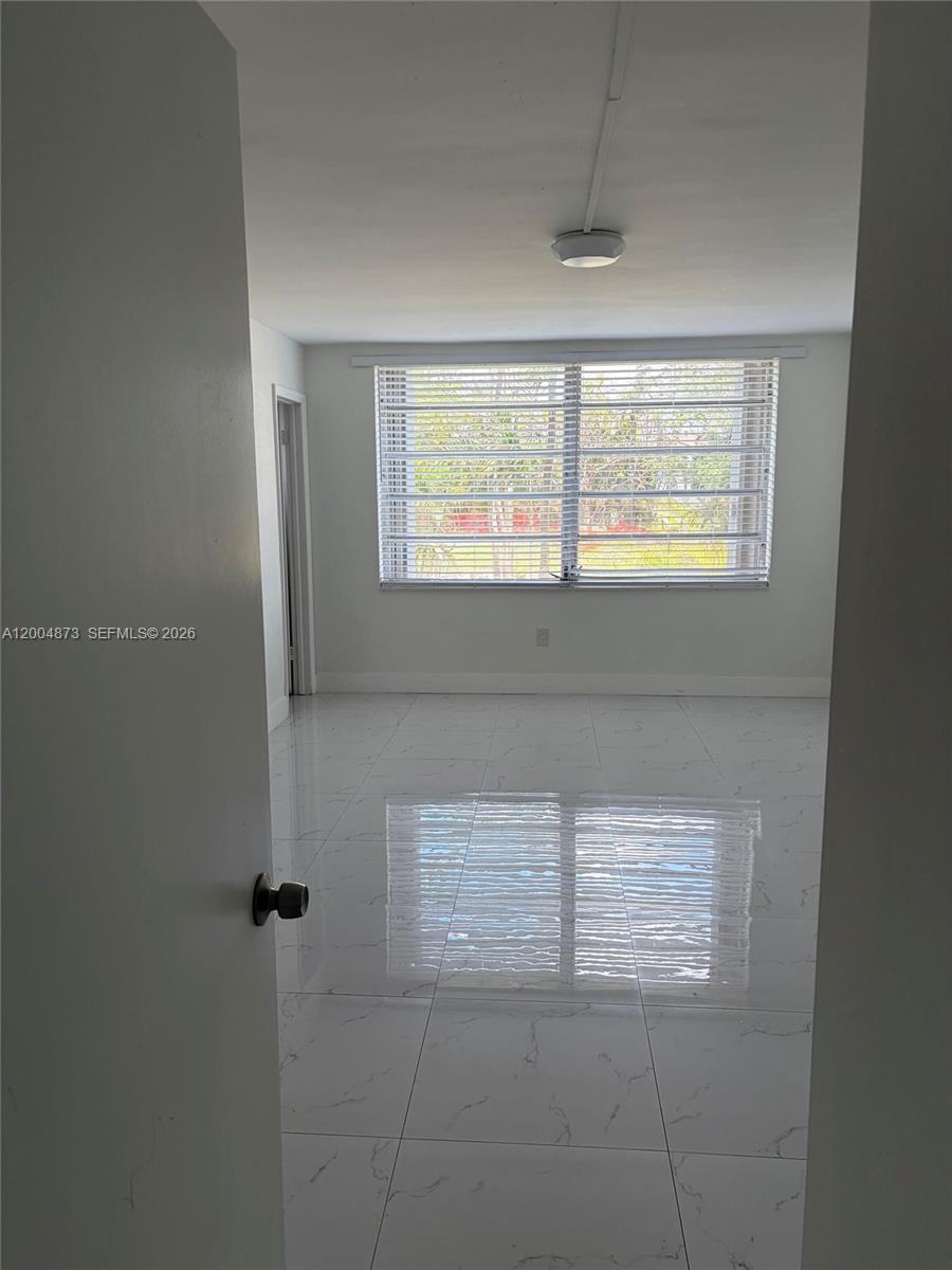 An empty room features white marble-look tile flooring, white walls, and a large window — 1401 NE 191st St # D201, Miami FL 33179, MIAMI, FL