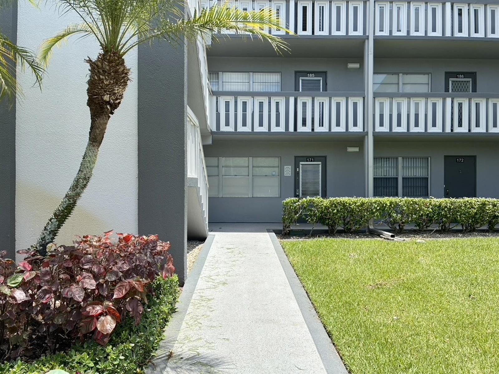 A concrete walkway leads toward the entrance of a two-story apartment building with gray siding — 171 Suffolk F # 171, Boca Raton FL 33434, BOCA RATON, FL