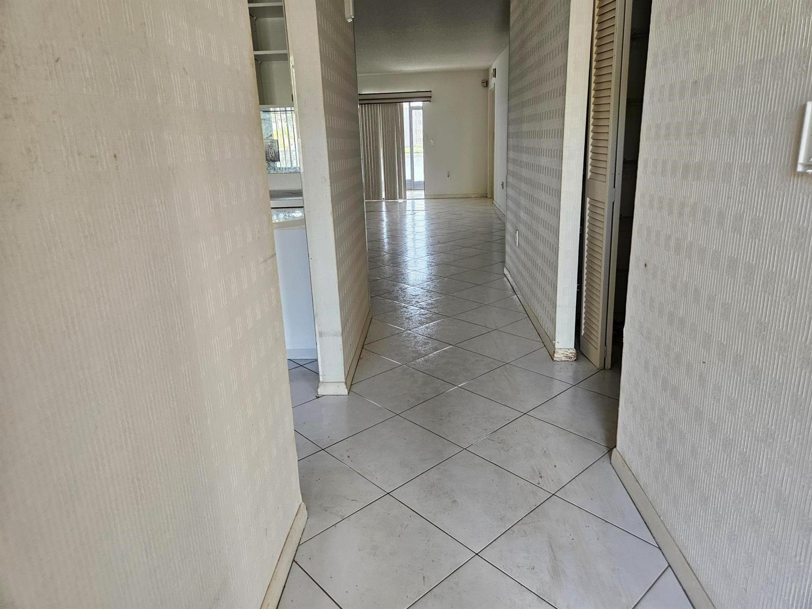 A hallway with light-colored tile flooring leads toward a living area with sliding glass doors — 6065 Pointe Regal Circle # 105, Delray Beach FL 33484, DELRAY BEACH, FL