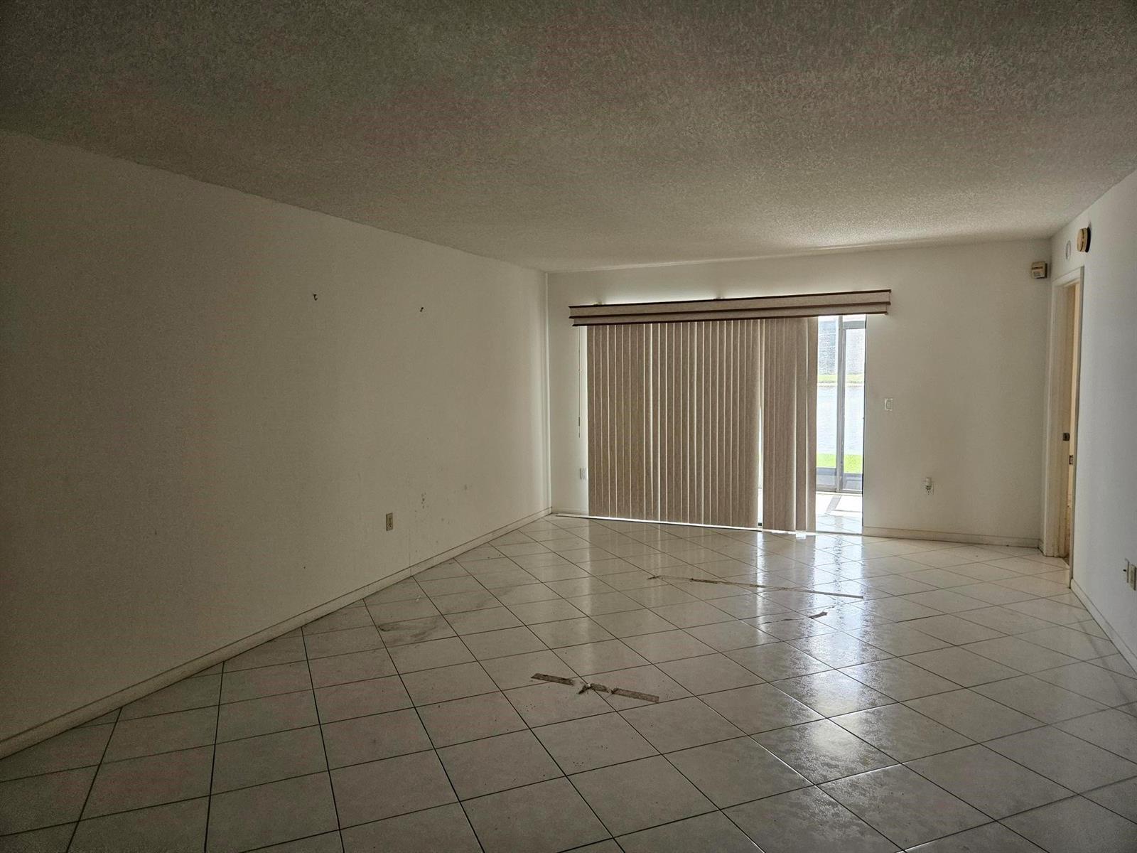 An empty living room features white walls, light-colored tile flooring, and a sliding glass door — 6065 Pointe Regal Circle # 105, Delray Beach FL 33484, DELRAY BEACH, FL