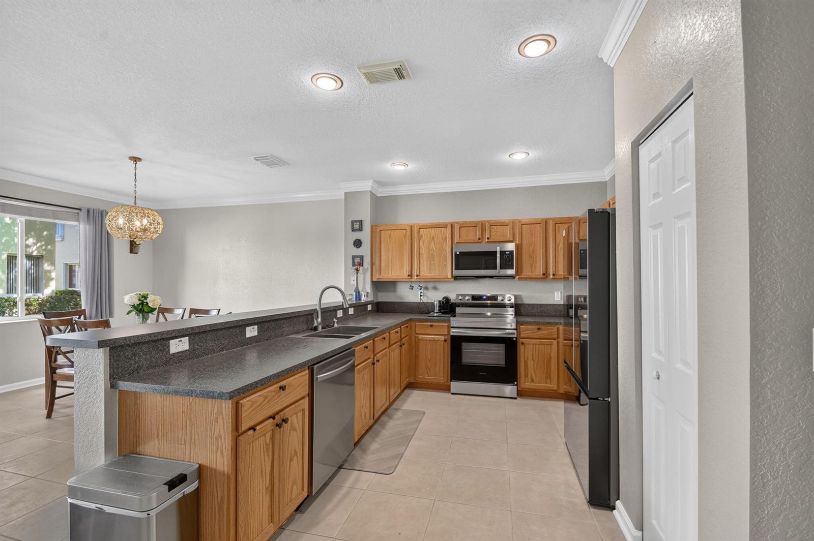A kitchen with oak cabinets, dark countertops, and stainless steel appliances opens to a dining area — 3117 N Evergreen Circle, Boynton Beach FL 33426, BOYNTON BEACH, FL