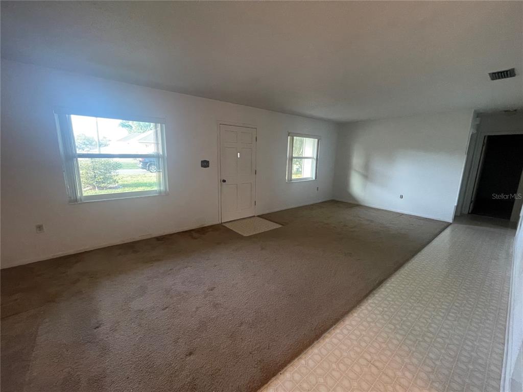An empty living room features brown carpeting, white walls, two windows, and a front door — 23 LONESOME PINE TRAIL, YALAHA, FL