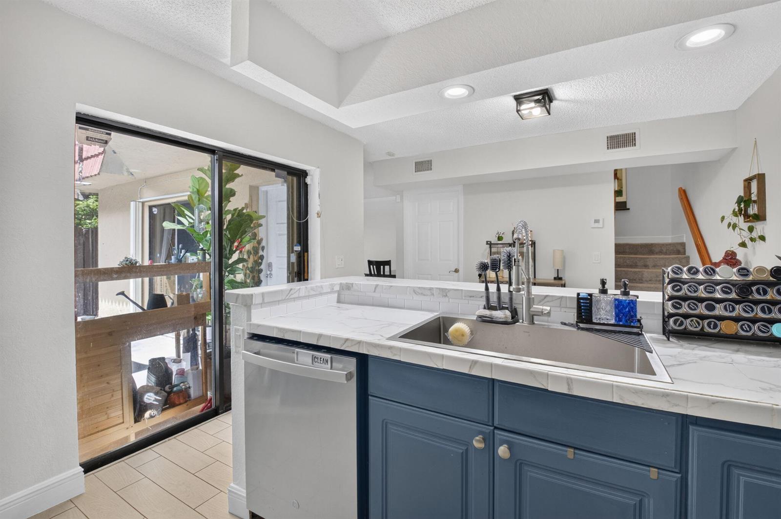A kitchen sink and dishwasher with blue cabinets and marble-look countertops face a sliding glass door — 22976 Oxford Place # C, Boca Raton FL 33433, BOCA RATON, FL