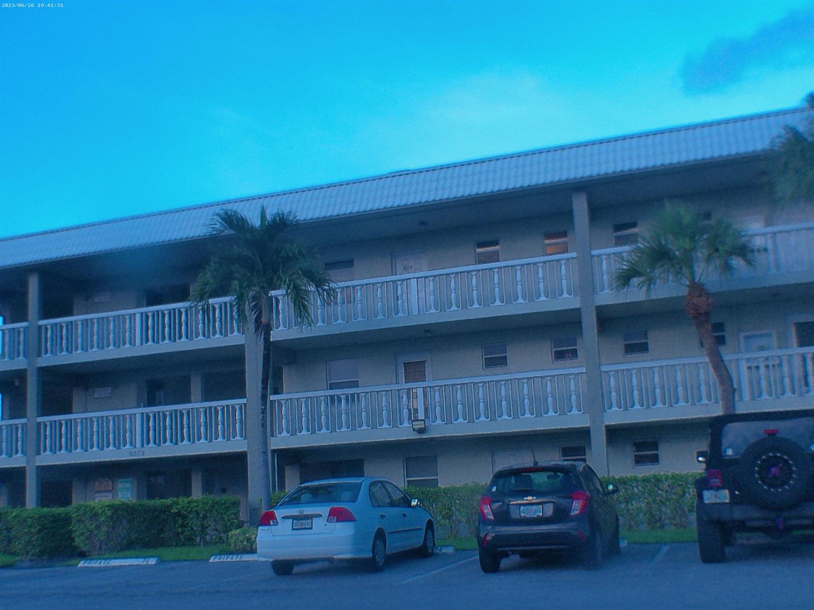 A three-story apartment building with balconies, palm trees, and parked cars in the foreground — 9872 Marina Boulevard # 1434, Boca Raton FL 33433, BOCA RATON, FL
