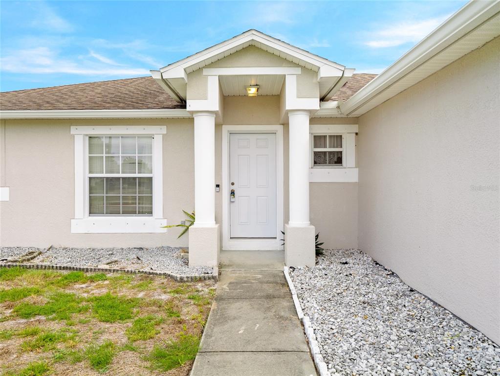 A beige house exterior features a white front door under a covered porch with columns — 867 NE FAIRHAVEN, PALM BAY, FL