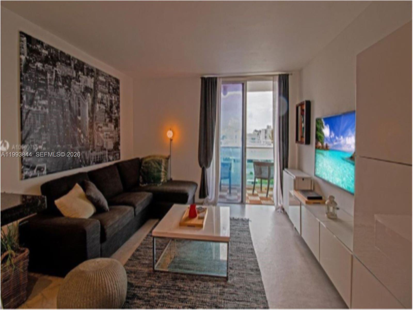 A living room features a dark sectional sofa, a coffee table, and a wall-mounted television — 1035 West Ave # 802, Miami Beach FL 33139, MIAMI BEACH, FL
