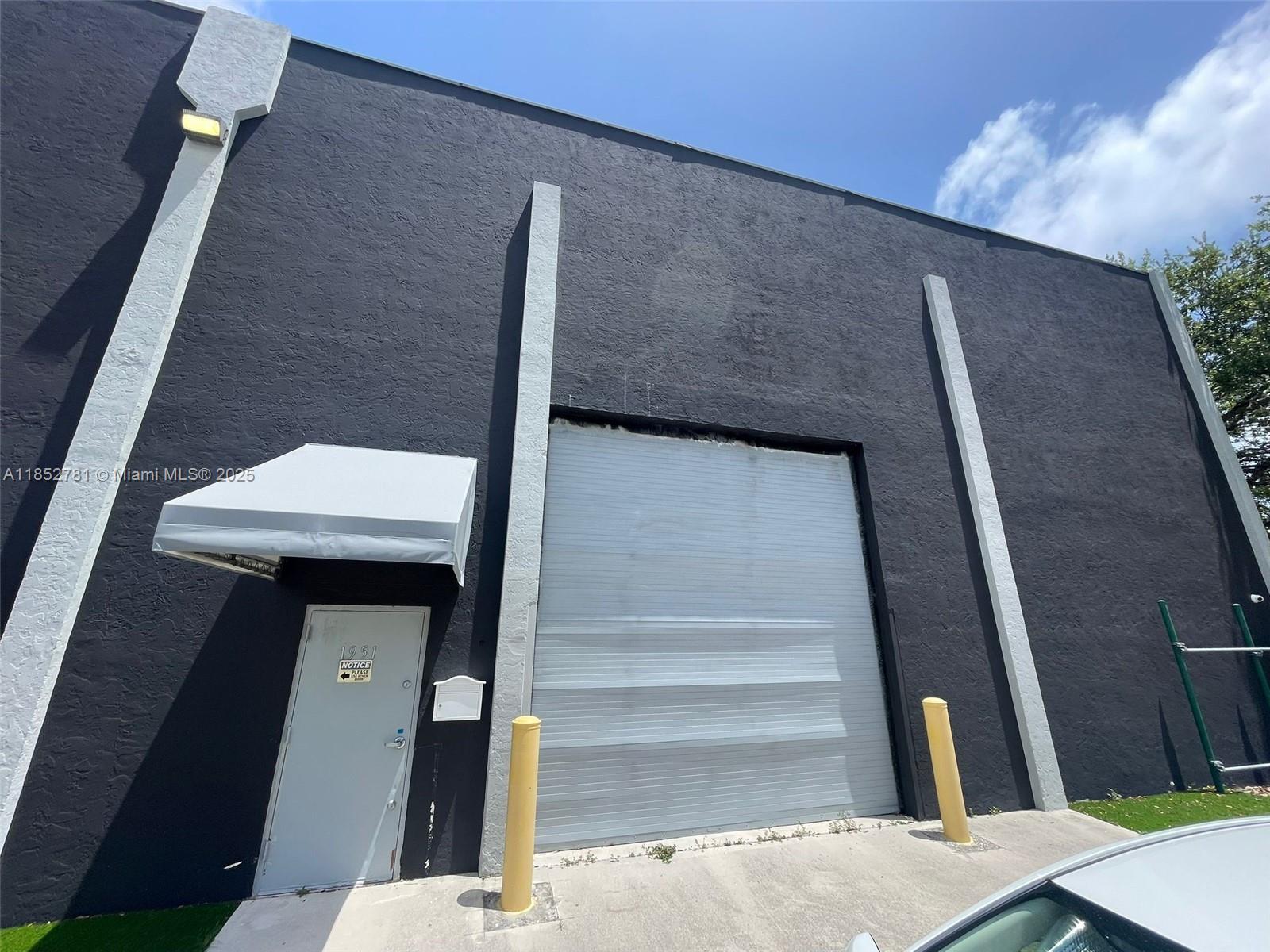 A black industrial building exterior features a gray roll-up door, a side entrance, and bollards — 1951 Grant St, Hollywood FL 33020, HOLLYWOOD, FL