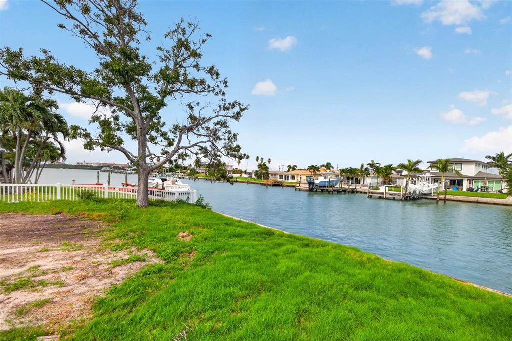 A grassy waterfront lot features a large tree, a white fence, and a canal view — 643 NORMANDY ROAD, MADEIRA BEACH, FL