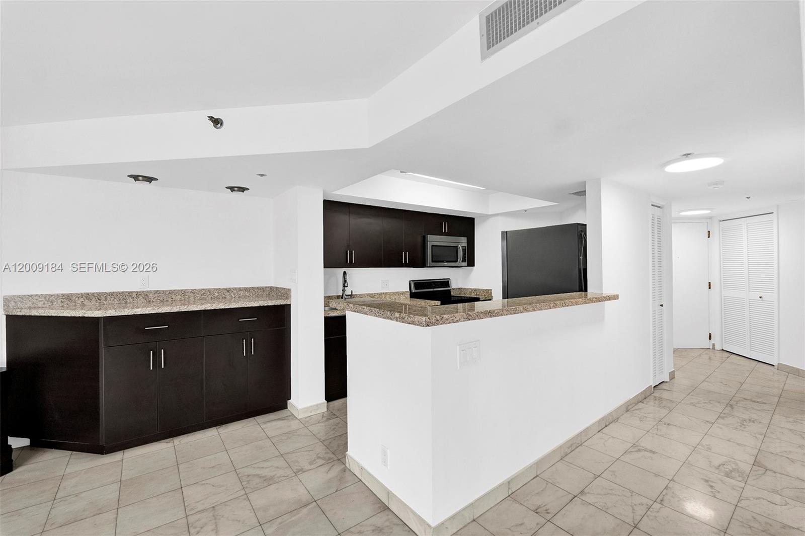 A kitchen features dark cabinetry, granite countertops, and light tile flooring in an open layout — 300 Three Islands Blvd # 305, Hallandale Beach FL 33009, HALLANDALE BEACH, FL