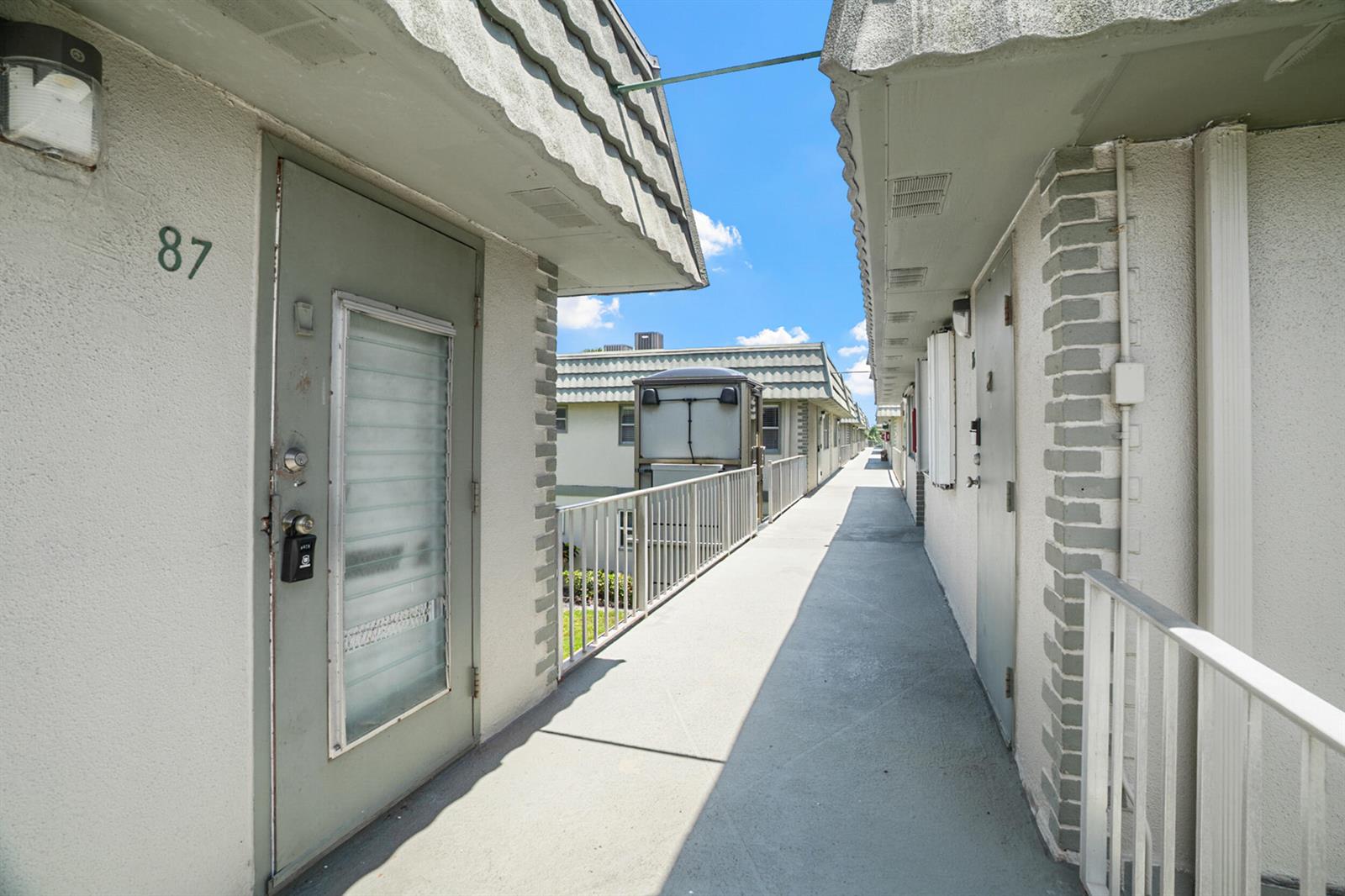 An outdoor walkway connects apartment units with gray doors and a metal stair lift — 87 Brittany B Avenue # B, Delray Beach FL 33446, DELRAY BEACH, FL