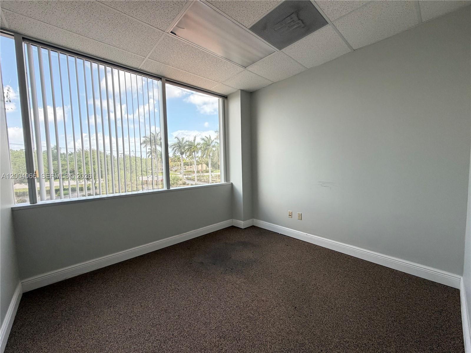 An empty office with gray walls, brown carpet, and large windows overlooking palm trees — 4400 W Sample Rd # 230, Coconut Creek FL 33073, COCONUT CREEK, FL
