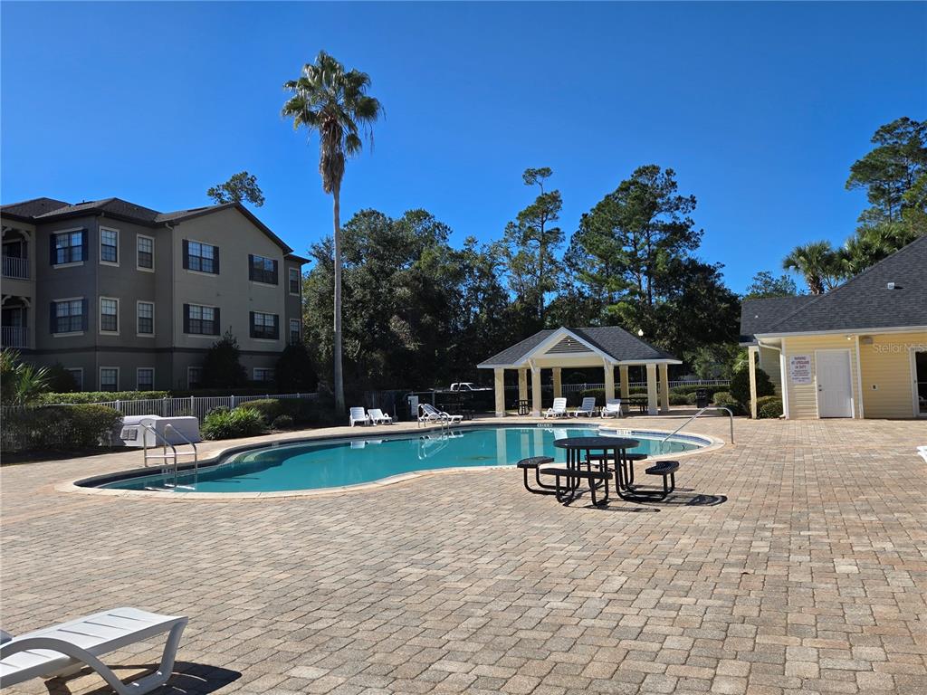 A swimming pool with a gazebo and patio furniture sits beside a multi-story apartment building — 4872 NW 42ND ROAD UNIT 103, GAINESVILLE, FL
