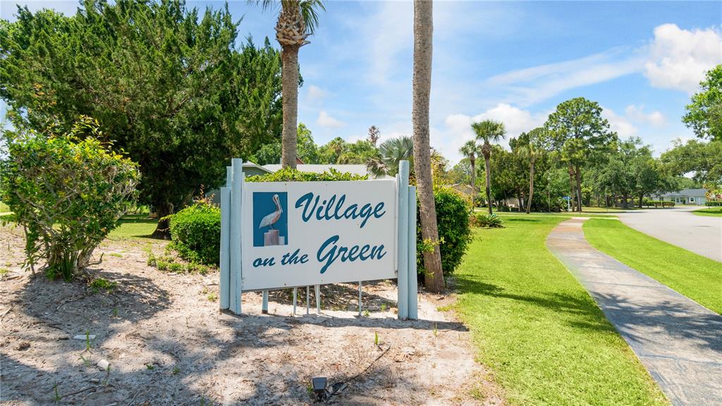 A sign for Village on the Green stands in a grassy area with trees — 108 GREBE COURT, DAYTONA BEACH, FL