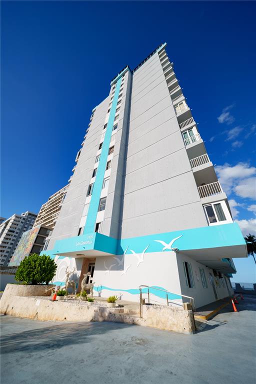 A low-angle exterior view of a tall, light-colored apartment building with blue accents — 3409 ISLA VERDE AVENUE UNIT 701, CAROLINA, PR