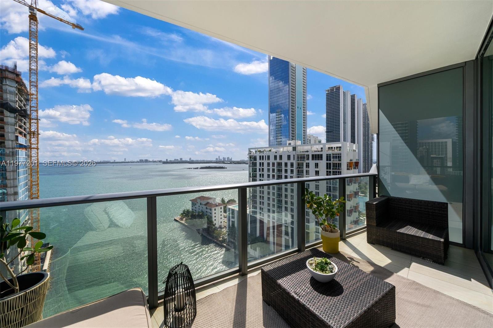 A balcony with wicker furniture overlooks a bay, nearby high-rise buildings, and a construction crane — 460 NE 28th St # 1901, Miami FL 33137, MIAMI, FL