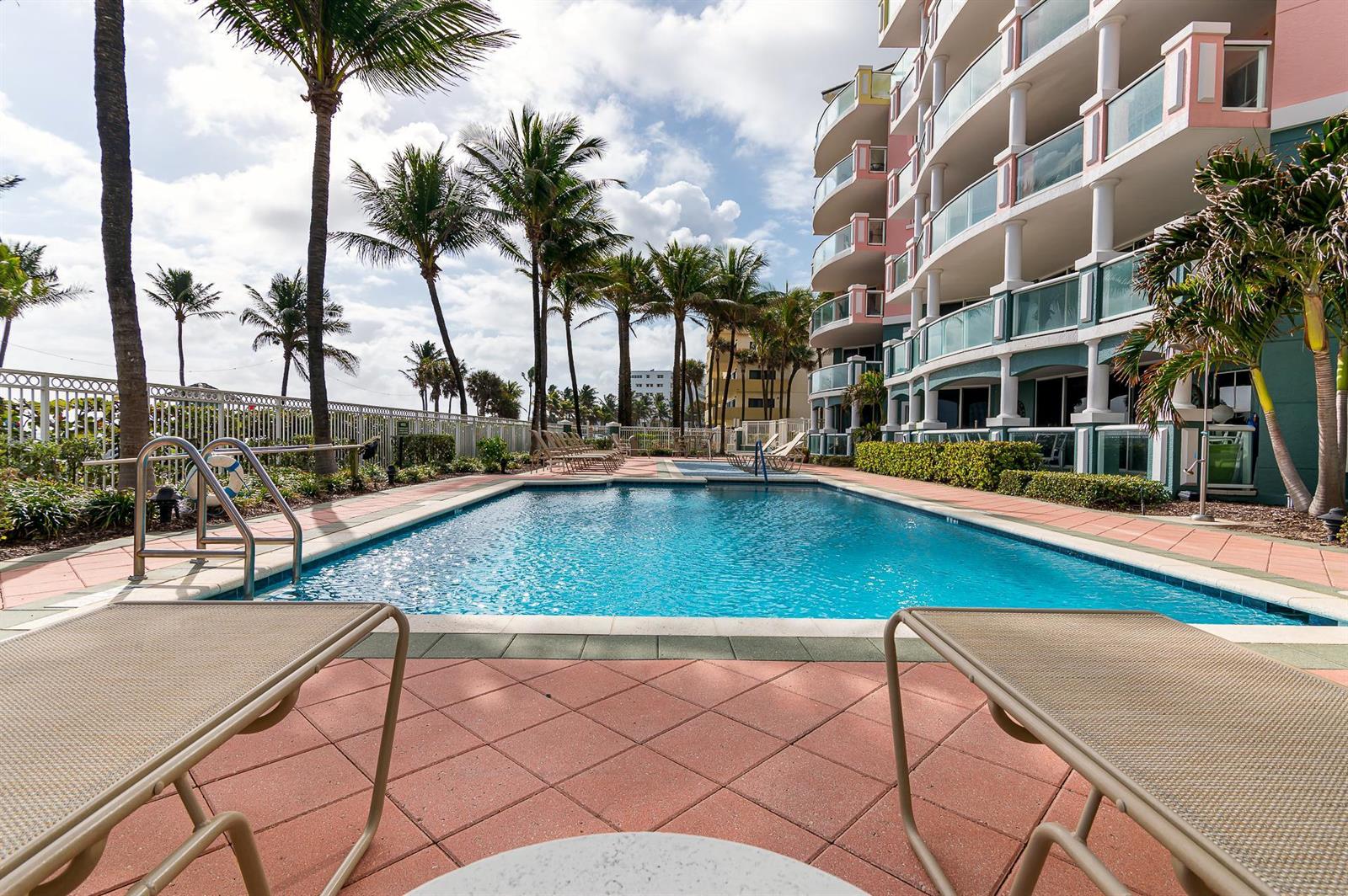 A swimming pool sits on a tiled deck next to a multi-story building and palm trees — 2051 SE 3rd Street # 101, Deerfield Beach FL 33441, DEERFIELD BEACH, FL
