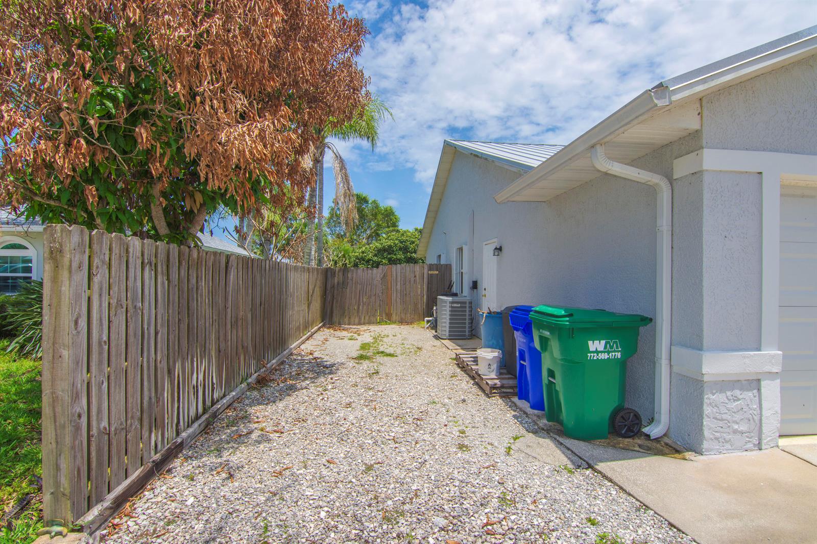 A gravel side yard features a wooden fence, trash bins, and an air conditioning unit — 474 Arbor Street, Sebastian FL 32958, SEBASTIAN, FL