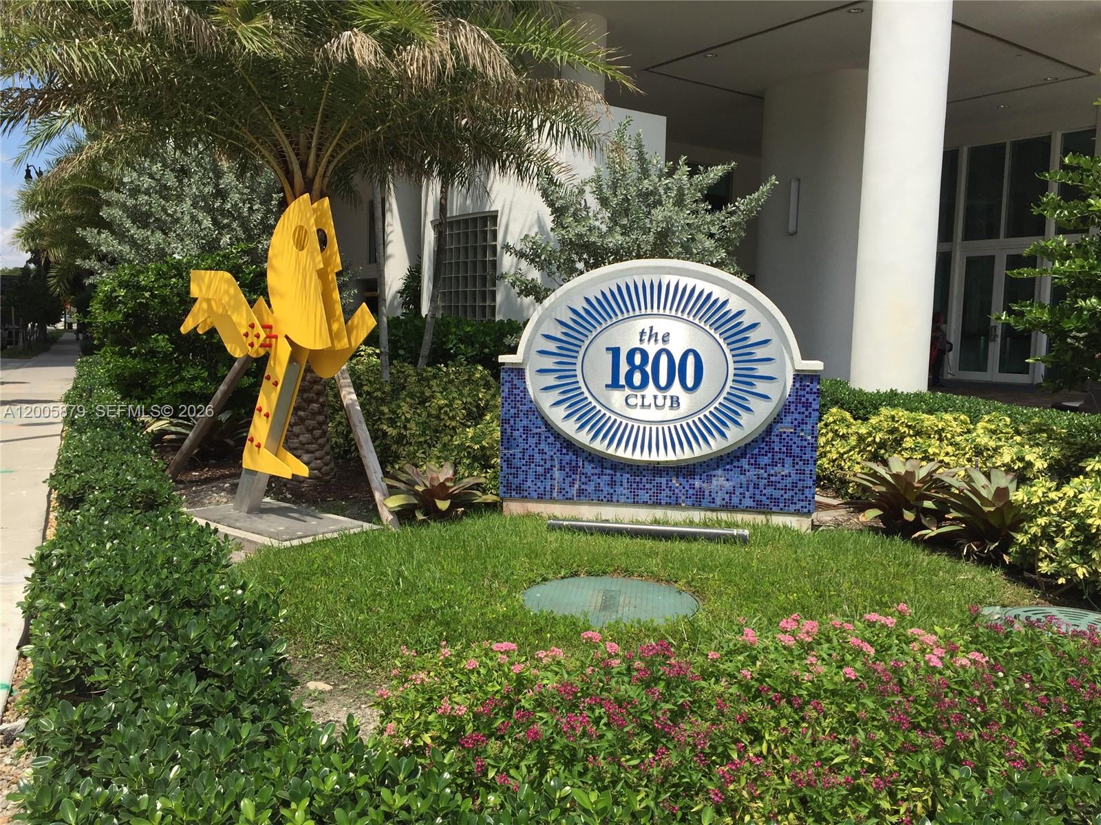 A sign for The 1800 Club stands in a landscaped garden with a yellow sculpture — 1800 N Bayshore Dr # 1506, Miami FL 33132, MIAMI, FL