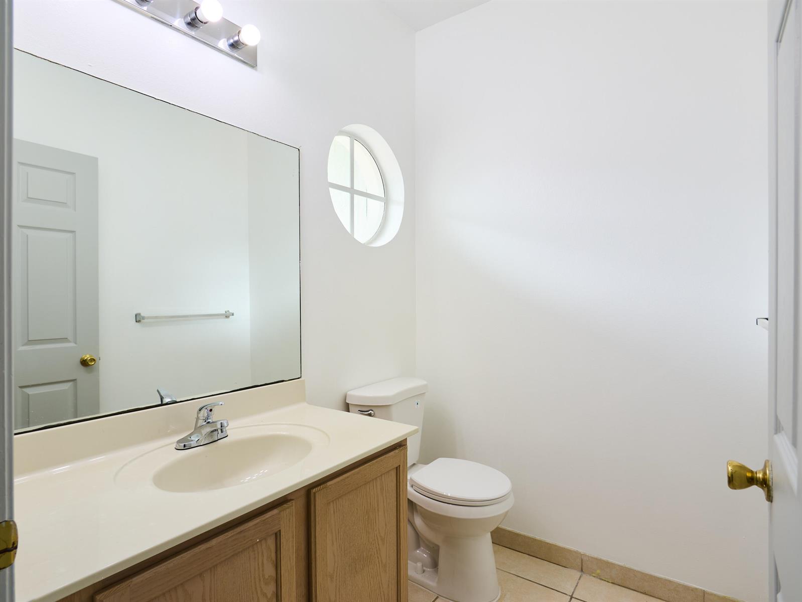 A bathroom features a wooden vanity, a large mirror, a toilet, and a circular window — 2052 Oakhurst Way, Riviera Beach FL 33404, RIVIERA BEACH, FL